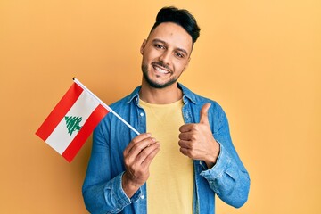 Young arab man holding lebanon flag smiling happy and positive, thumb up doing excellent and...
