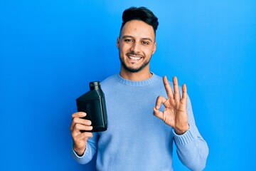 Young arab man holding motor oil bottle doing ok sign with fingers, smiling friendly gesturing excellent symbol