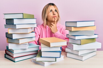 Young caucasian woman sitting on the table with books thinking concentrated about doubt with finger on chin and looking up wondering