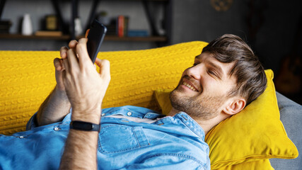 Relaxed smiling young man texting on cellphone lying on sofa at home, side view. Cheerful guy enjoying exchanging text messages with friends or beloved one