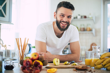 Handsome young sporty smiling man in the kitchen is preparing vegan healthy fruits salad and smoothie in a good mood
