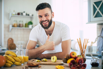 Handsome young sporty smiling man in the kitchen is preparing vegan healthy fruits salad and smoothie in a good mood