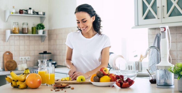 Cute beautiful and happy young brunette woman in the kitchen at home is preparing fruit vegan salad or a healthy smoothie and having fun