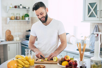 Handsome young sporty smiling man in the kitchen is preparing vegan healthy fruits salad and...