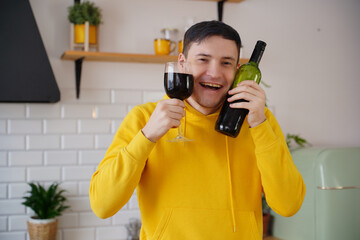 Relaxed young man poses with bottle and glass of red wine in kitchen. Adult happy guy presses alcohol to face.