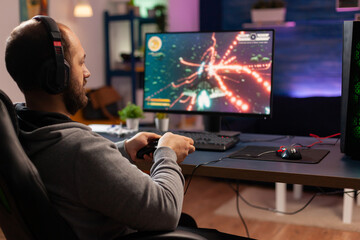 Concentrated gamer playing virtual game on powerful computer at home with professional headphones. Digital gamer using joystick for shooter space gaming competition late at night in living room