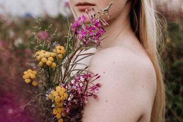 Young woman with naked sholders holding bouquet of yellow and purple wild flowers standing in green...