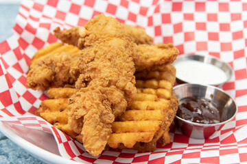 Chicken tenders breaded and cooked to perfection sitting on top of a pile of waffle french fries along with dipping sauces.