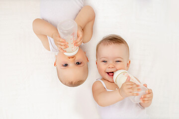twin babies boy and girl with a bottle of milk on a white bed at home, baby food concept, place for text - obrazy, fototapety, plakaty