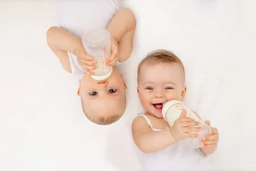 Fototapeten twin babies boy and girl with a bottle of milk on a white bed at home, baby food concept, place for text © Any Grant