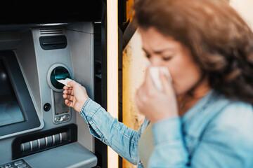 Young woman coughing and sneezing while using ATM machine to withdraw cash. Allergy, flu, virus concept.