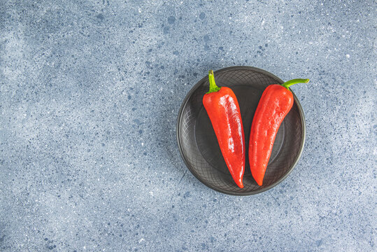 Fresh raw sweet red pepper kapi with water drops in black ceramic plate, two peppers on gray table surface.