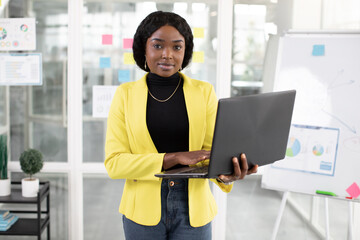 Pretty young professioanl female African American office worker, dressed in casual hipster outfit, posing to camera with laptop pc, standing in modern light office with flipchart on the background.