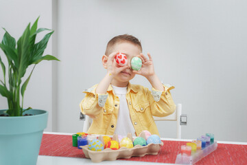 Little boy holds Easter eggs by the eyes, Easter holiday concept. Close-up portrait of a child in home interior. Decorated Easter eggs