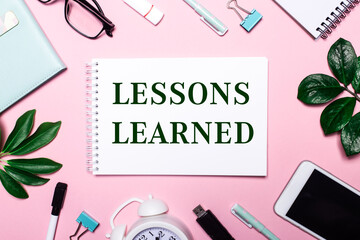 Fototapeta na wymiar LESSONS LEARNED is written in a white notebook on a pink background surrounded by business accessories and green leaves.