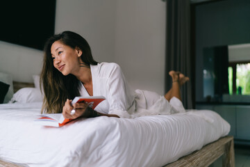 Dreamy Asian female reading book in bedroom