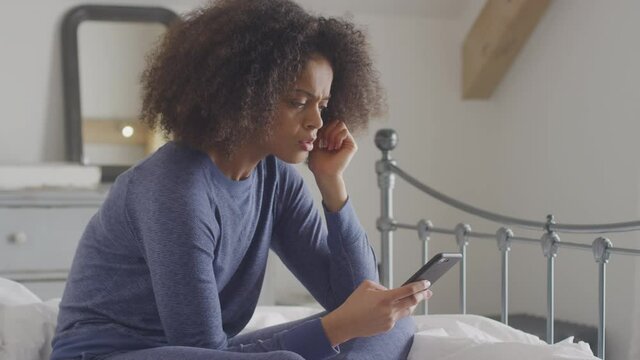 Unhappy woman wearing pyjamas sitting on bed with mobile phone being bullied online via social media - shot in slow motion
