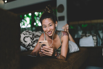 Smiling ethnic woman using smartphone and earphones on sofa in flat