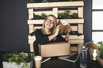 pretty young blonde woman with brown parcels is sitting in front of laptop in a very modern, ecological, sustainable office and is happy about her online order