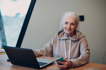 Aged elderly woman with laptop and mobile phone working on computer at table