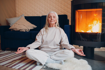 Portrait of senior woman sitting in lotus position indoors with fireplace. Yoga and meditation zen...