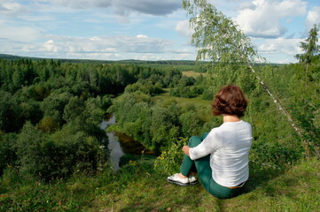 a woman sits on the steep bank of the river and looks into the distance