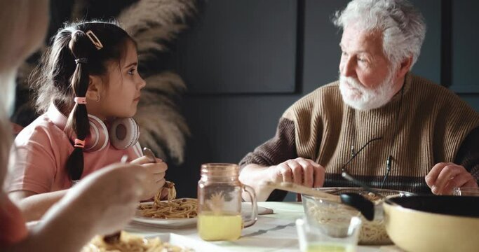 Grandpa talks to little girl granddaughter at table while having lunch, happy family eating spaghetti together. A little girl with headphones listens to her grandfather as she talks
