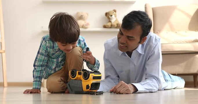 Caring young indian ethnicity father teaching cute little kid son repairing toy car with screwdriver, playing together in living room, lying on warm floor, enjoying weekend leisure domestic activity.