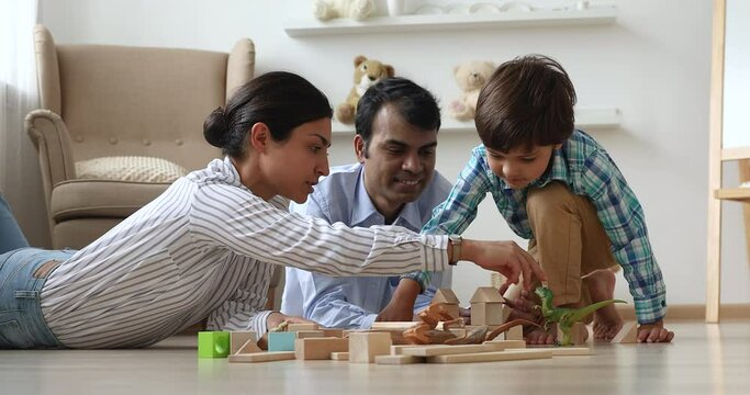Happy young mixed race indian family couple playing toys with small adorable preschool child son, sitting together on warm wooden floor, enjoying spending free weekend time together at home.