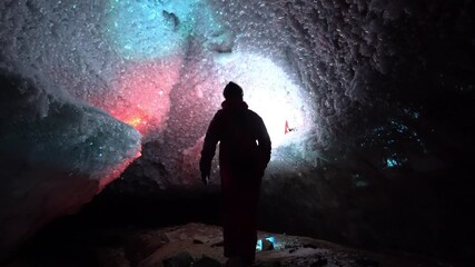 A guy in an ice cave with a lantern light. The caver descended into the ice cave. Snow stalactites and ice walls. In some places there are stones. Colored lantern beams. Bogdanovich Glacier, Almaty