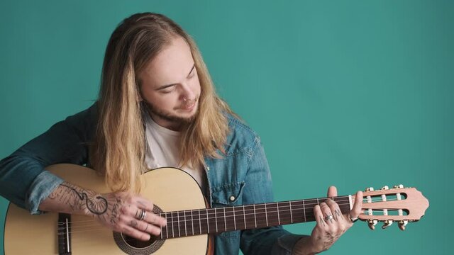 Young long haired blond man playing on acoustic guitar preparing to perform over blue background. Music concept