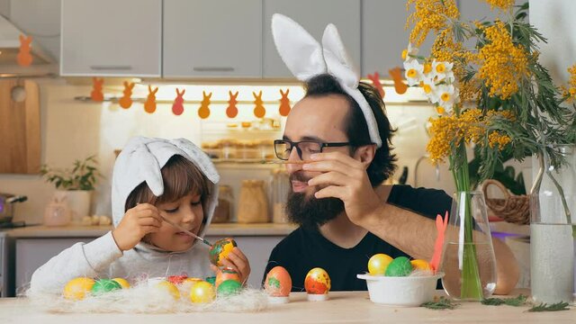 Happy Easter. Father in rabbit ears and young son draw on painted eggs at the table, at home. Funny family prepares for holiday