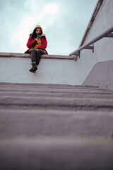 Young woman with mobile phone sitting on concrete parapet. Brunette in yellow hoodie and red jacket browsing smartphone in cloudy weather.