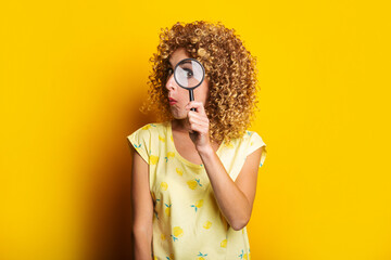 surprised curly young woman looks through a magnifying glass on a yellow background.
