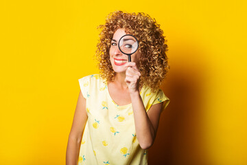 smiling curly young woman looking through a magnifying glass on a yellow background.