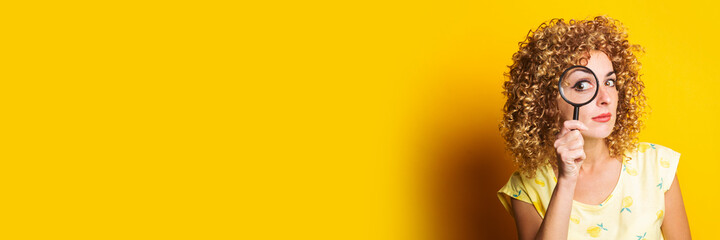 curly young woman looks through a magnifying glass on a yellow background. Banner.