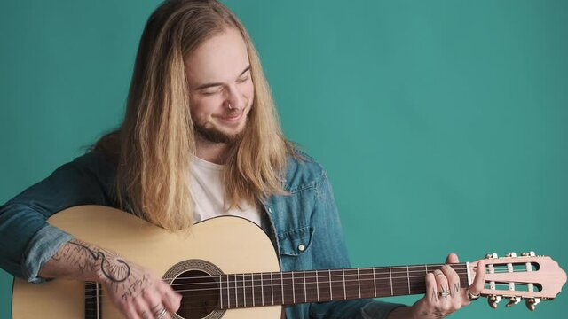 Young long haired blond man playing on acoustic guitar enjoying it isolated on blue background. Music concept