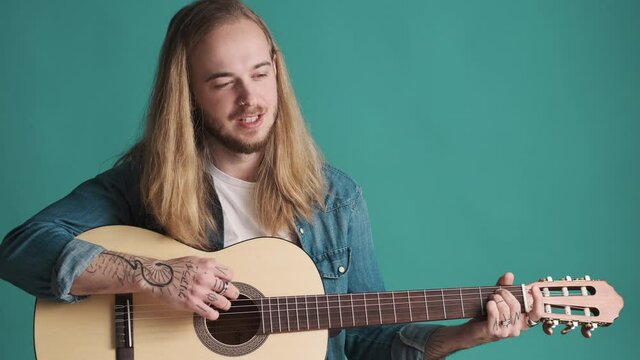 Young long haired blond man playing on acoustic guitar and singing new song over blue background. Music concept