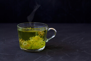 hot linden flowers tea.Cup of hot herbal tea with linden fresh flowers on a black table.Healthy lifestyle.glass cup of tea with green leaves on a black background.hot drinks.copy space.