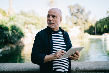 Contemplative Caucaisan man 60 years old holding digital tablet thoughtful looking away during...