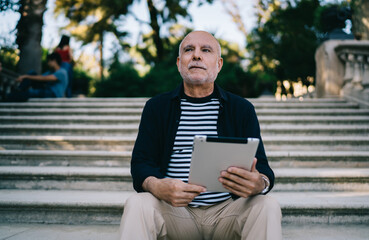 Obraz premium Pensive male tourist resting at ancient stairs in city feeling pondering on travel lifestyle, thoughtful senior with modern digital technology for make booking thinking about vacations outdoors