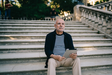 Fototapeta premium Pensive male tourist resting at ancient stairs in city feeling pondering on travel lifestyle, contemplative senior with modern digital technology for make booking thinking about vacations outdoors