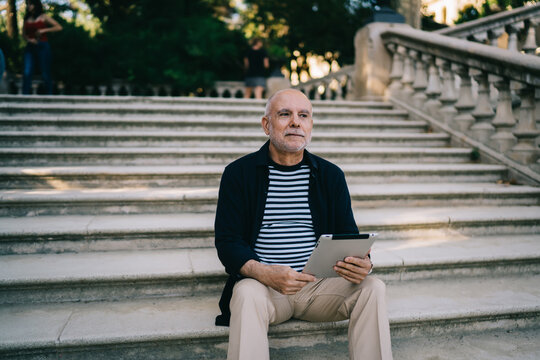 Pensive male tourist resting at ancient stairs in city feeling pondering on travel lifestyle, contemplative senior with modern digital technology for make booking thinking about vacations outdoors - Powered by Adobe
