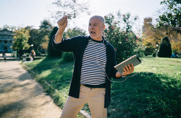 Emotional young in heart male pensioner with digital tablet having fun at public place enjoying free time, senior man in earphones listening energetic music from touch pad and dancing rock'n'roll