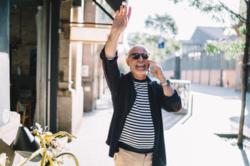 Fototapeta premium Funny male tourist discussing international vacations on retirement using roaming internet connection on cellphone device, cheerful elderly man waving and laughing in city while calling via mobile