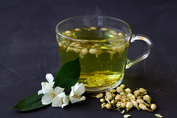 Close up Jasmine tea. Cup of hot herbal tea with jasmine fresh flowers on a black table. Healthy lifestyle. glass cup of green tea with dry leaves on a black background. Hot drinks.