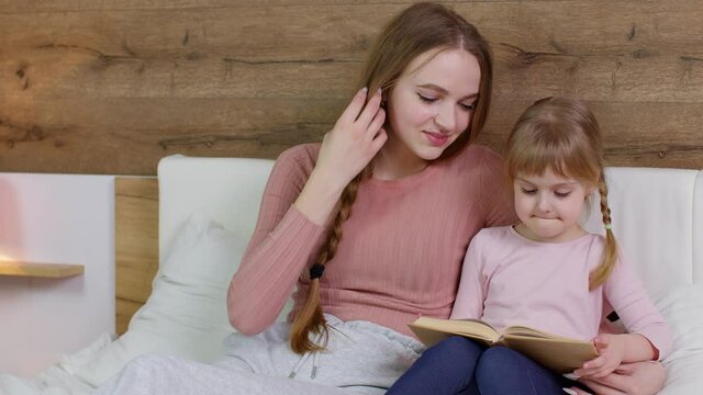 Young mother reading goodnight story book fairytale to child daughter kid girl in night bedroom
