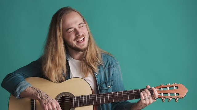 Young long haired blond bearded man playing on acoustic guitar and singing new song looking inspiring over colorful background. Music concept