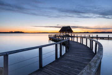 Fototapeta premium Swinging Pier construction and shelter with thatched roof at beautiful cool dawn before sunrise under cloudy sky at Lake Hemmelsdorf, Schleswig-Holstein, Northern Germany