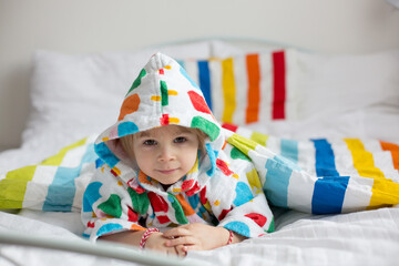 Happy toddler child, blond boy with colorful bathrobe, sitting in bed with stuffed toy after bath, smiling